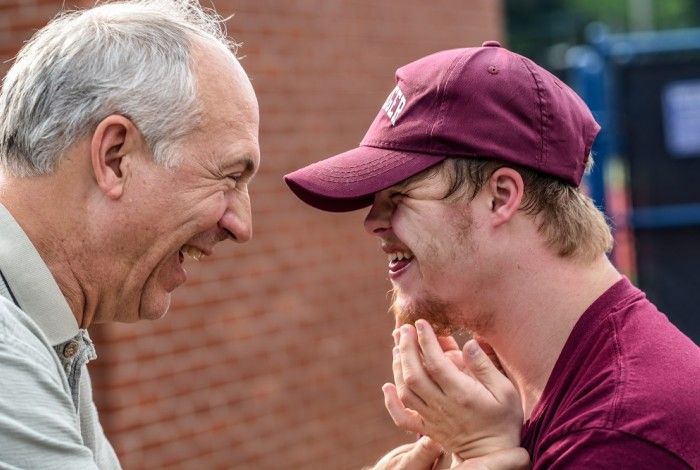 Two people smiling and engaging warmly outdoors, with one older man and one younger man wearing a maroon cap, sharing a joyful moment together.