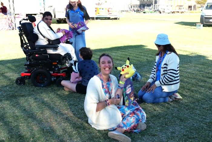A group of people gathered outdoors on grass. One person in a wheelchair is seated while two others are sitting nearby. A woman in a white outfit is smiling and holding a Pikachu plush toy. Another person is standing, facing the group.