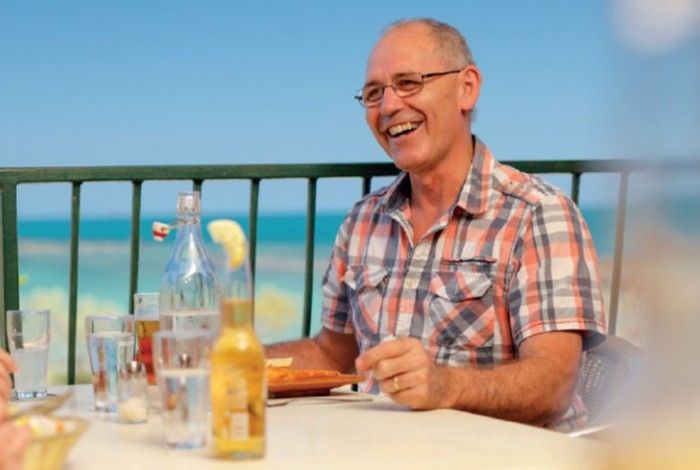 Smiling older man wearing glasses and a short-sleeved checked shirt, seated outdoors at a table with drinks and food, enjoying a meal with others beside a turquoise ocean backdrop.